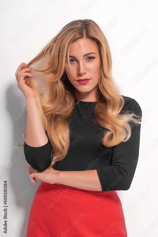 Blonde woman in a red skirt on the background of a white wall with a shadow.