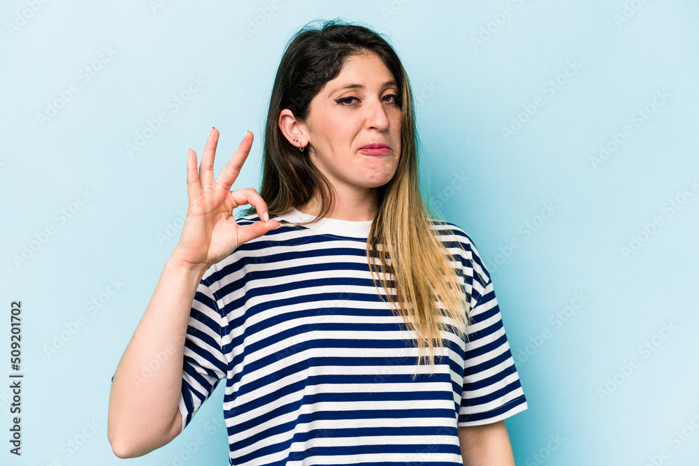Young caucasian woman isolated on blue background winks an eye and holds an okay gesture with hand. Young caucasian woman isolated on blue background winks an eye and holds an okay gesture with hand.