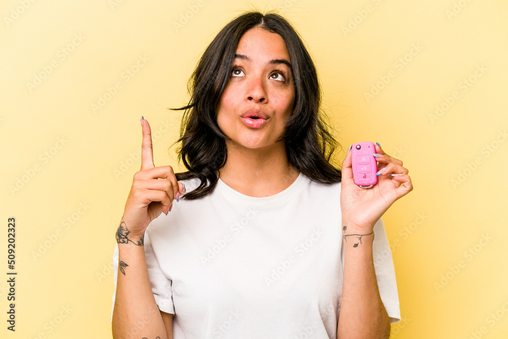 Young hispanic woman holding a keys car isolated on pink background pointing upside with opened mouth. Young hispanic woman holding a keys car isolated on pink background pointing upside with opened mouth.