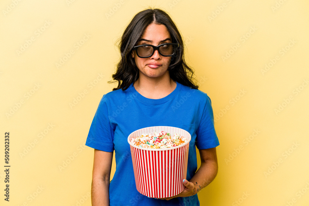 Young hispanic woman holding popcorn isolated on yellow background confused, feels doubtful and unsure.