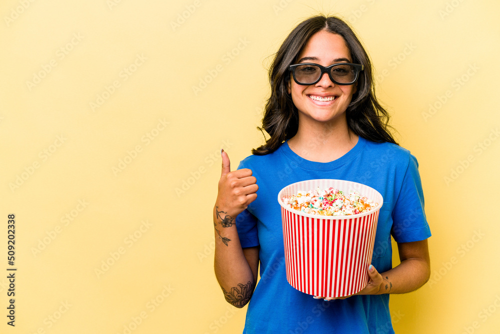 Young hispanic woman holding popcorn isolated on yellow background smiling and raising thumb up