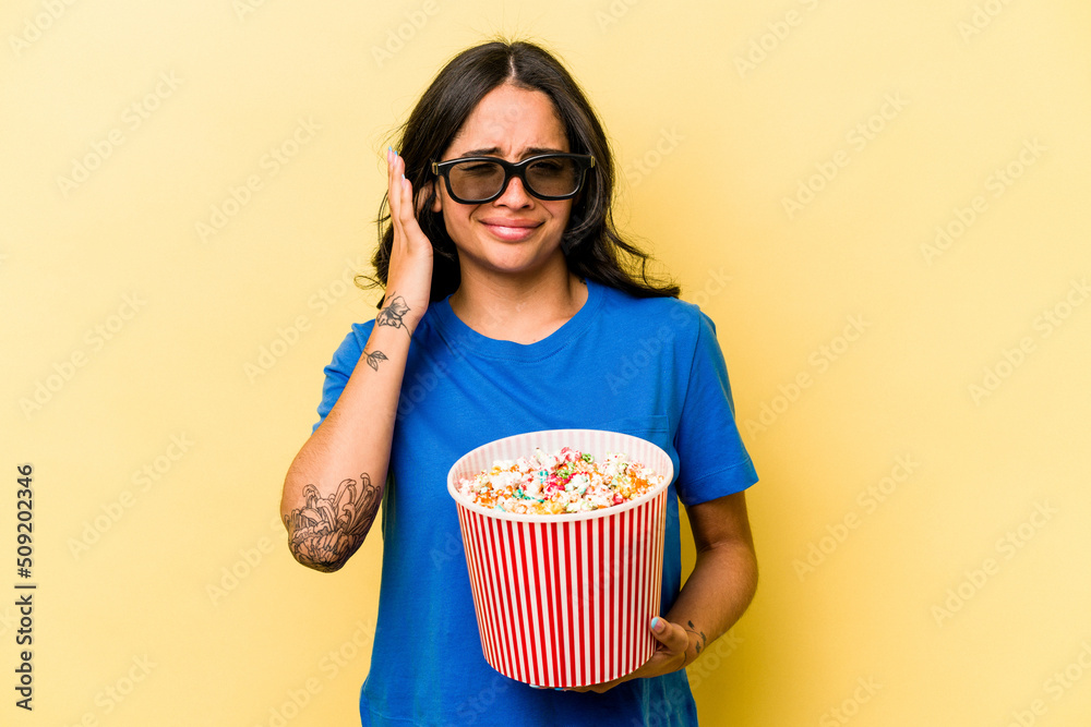 Young hispanic woman holding popcorn isolated on yellow background covering ears with hands.