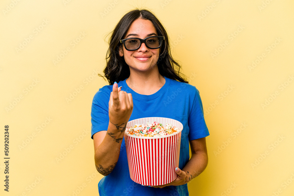 Young hispanic woman holding popcorn isolated on yellow background pointing with finger at you as if inviting come closer.