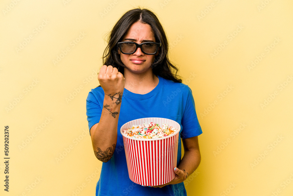 Young hispanic woman holding popcorn isolated on yellow background showing fist to camera, aggressive facial expression.