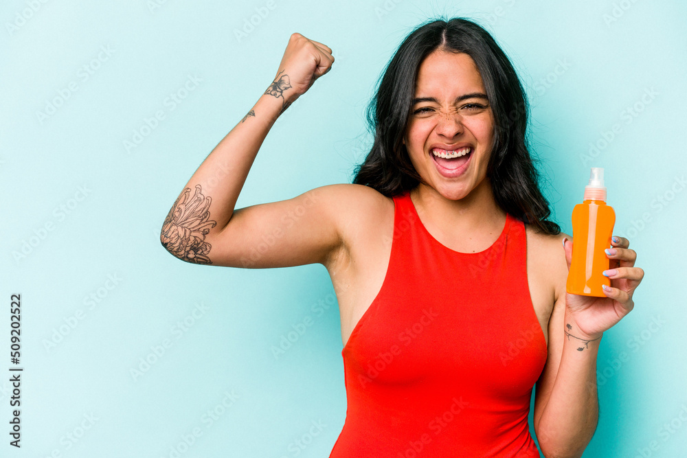Young hispanic woman holding sun cream isolated on blue background raising fist after a victory, winner concept. Young hispanic woman holding sun cream isolated on blue background raising fist after a victory, winner concept.