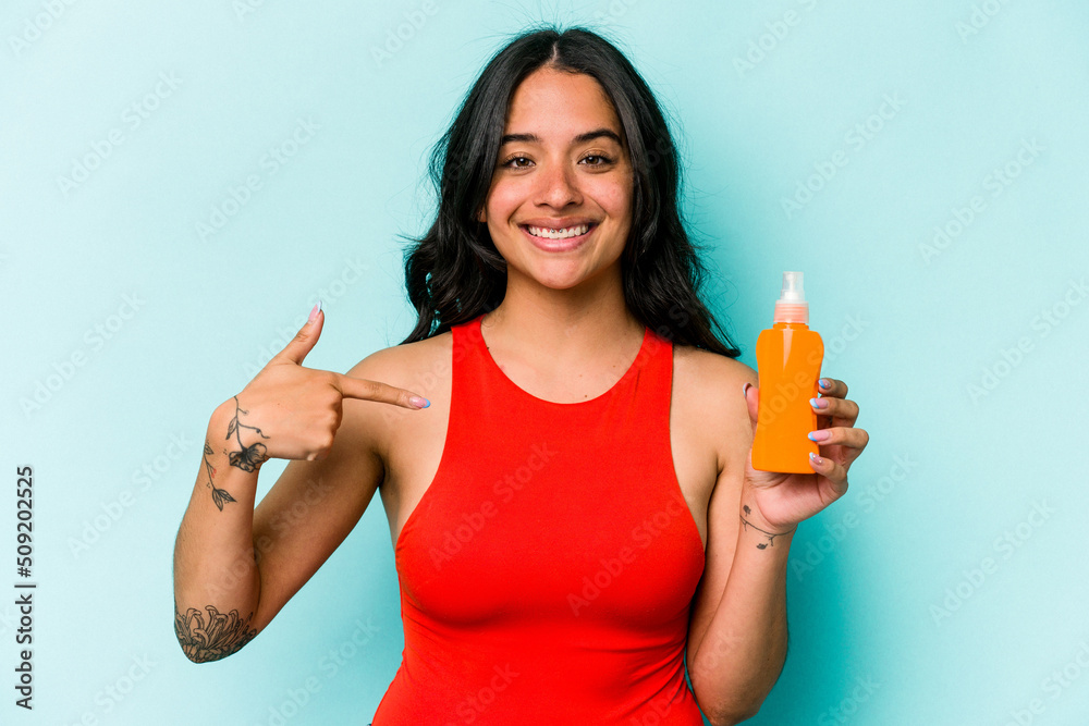 Young hispanic woman holding sun cream isolated on blue background person pointing by hand to a shirt copy space, proud and confident Young hispanic woman holding sun cream isolated on blue background person pointing by hand to a shirt copy space, proud and confident