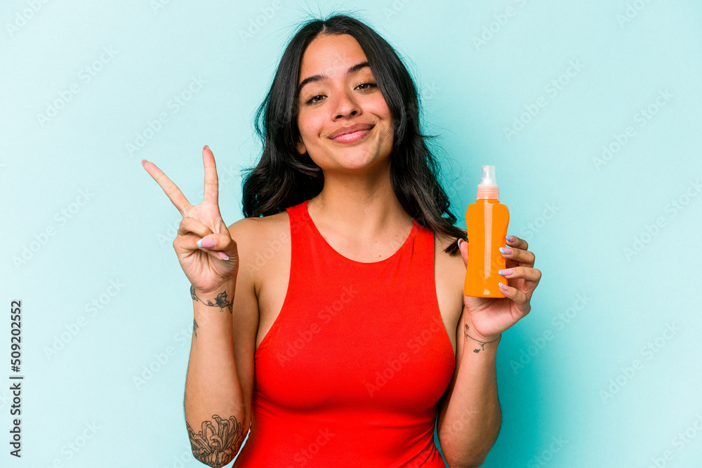 Young hispanic woman holding sun cream isolated on blue background showing number two with fingers. Young hispanic woman holding sun cream isolated on blue background showing number two with fingers.