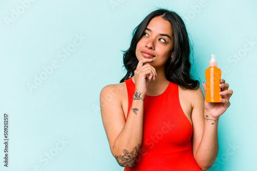 Young hispanic woman holding sun cream isolated on blue background looking sideways with doubtful and skeptical expression.