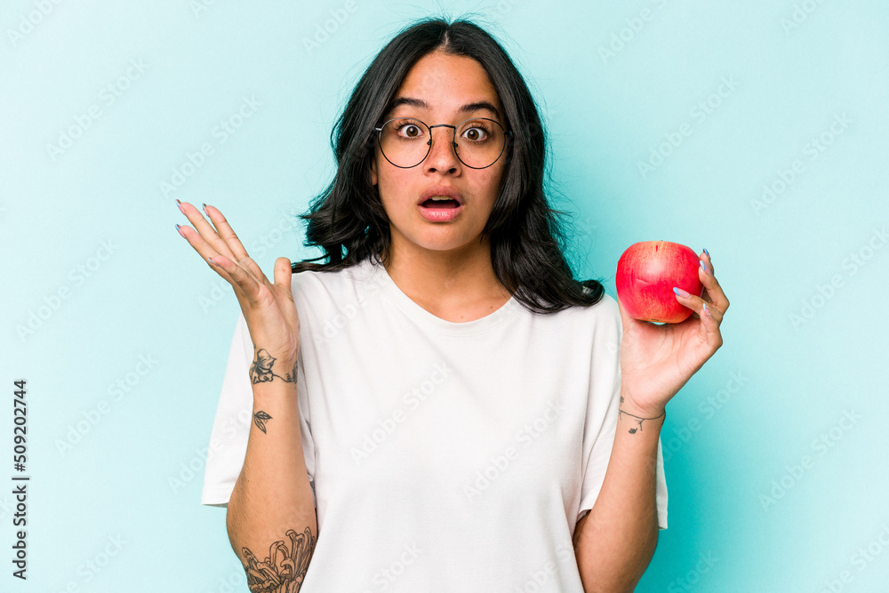 Young hispanic woman holding an apple isolated on blue background surprised and shocked.
