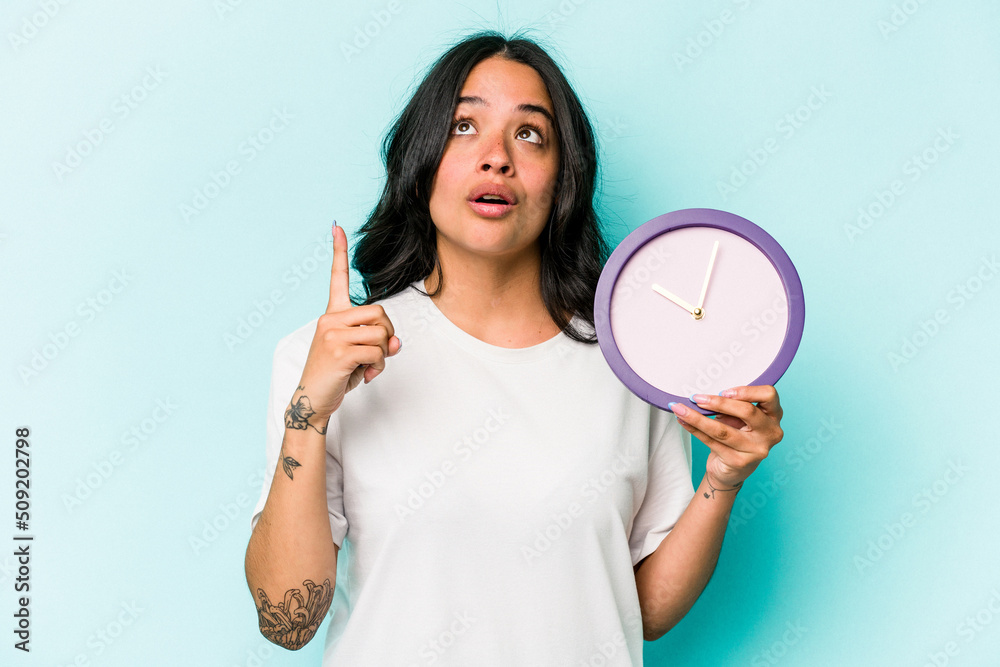Young hispanic woman holding a clock isolated on blue background pointing upside with opened mouth. Young hispanic woman holding a clock isolated on blue background pointing upside with opened mouth.