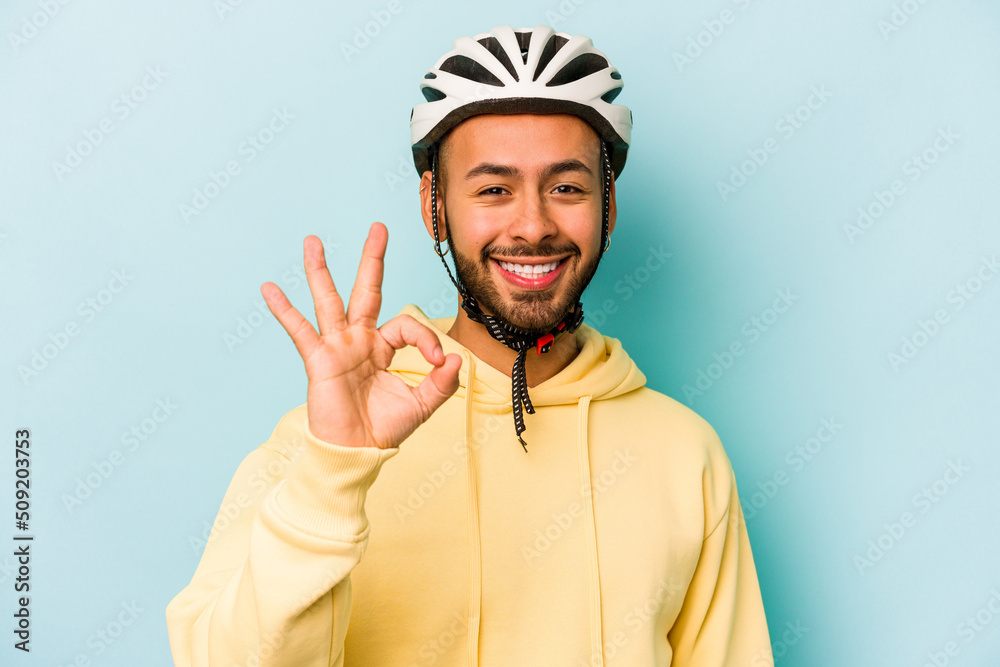 Young hispanic man wearing helmet isolated on blue background cheerful and confident showing ok gesture.