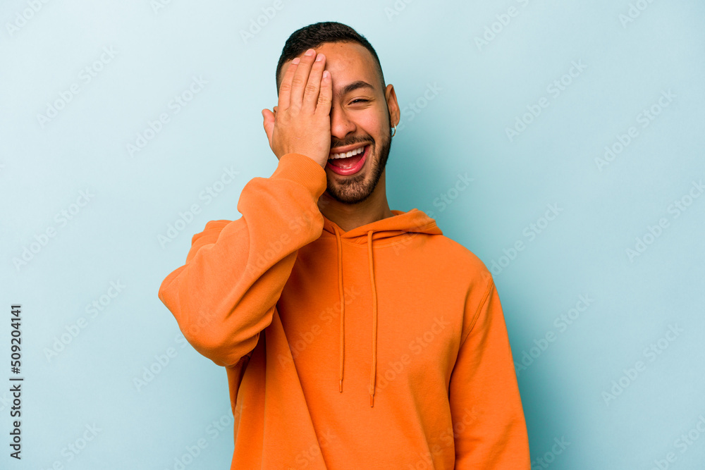 Young hispanic man isolated on blue background having fun covering half of face with palm.