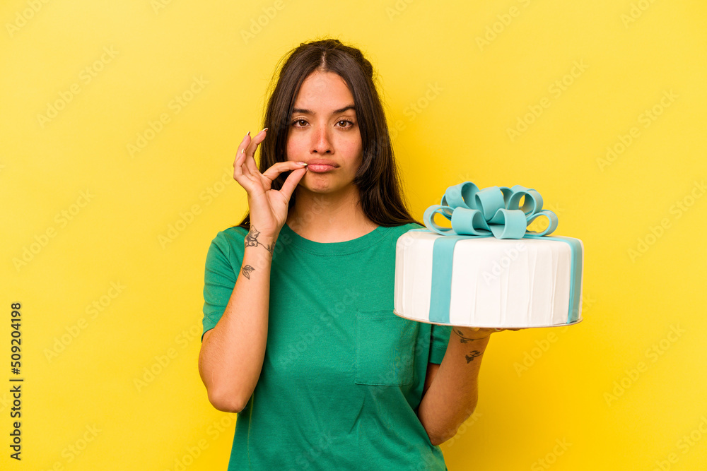 Young hispanic woman holding birthday cake isolated on yellow background with fingers on lips keeping a secret. Young hispanic woman holding birthday cake isolated on yellow background with fingers on lips keeping a secret.