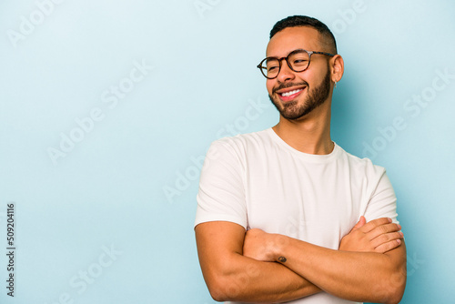 Young hispanic man isolated on blue background smiling confident with crossed arms.