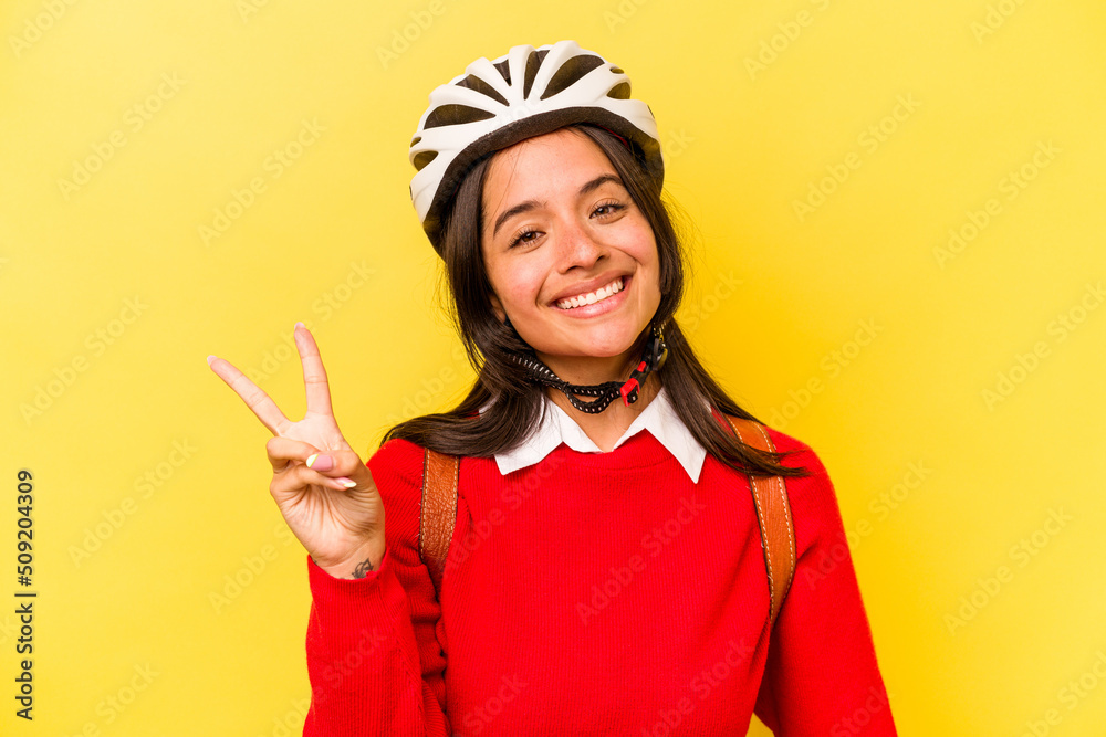 Young student hispanic woman wearing a bike helmet isolated on yellow background joyful and carefree showing a peace symbol with fingers. Young student hispanic woman wearing a bike helmet isolated on yellow background joyful and carefree showing a peace symbol with fingers.