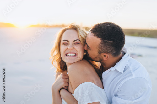 A tanned caucasian bearded guy in a white shirt kisses a young beautiful cheerful smiling blonde against the backdrop of the setting sun. Desert, sandy beach, rest and relaxation. Honeymoon concept.