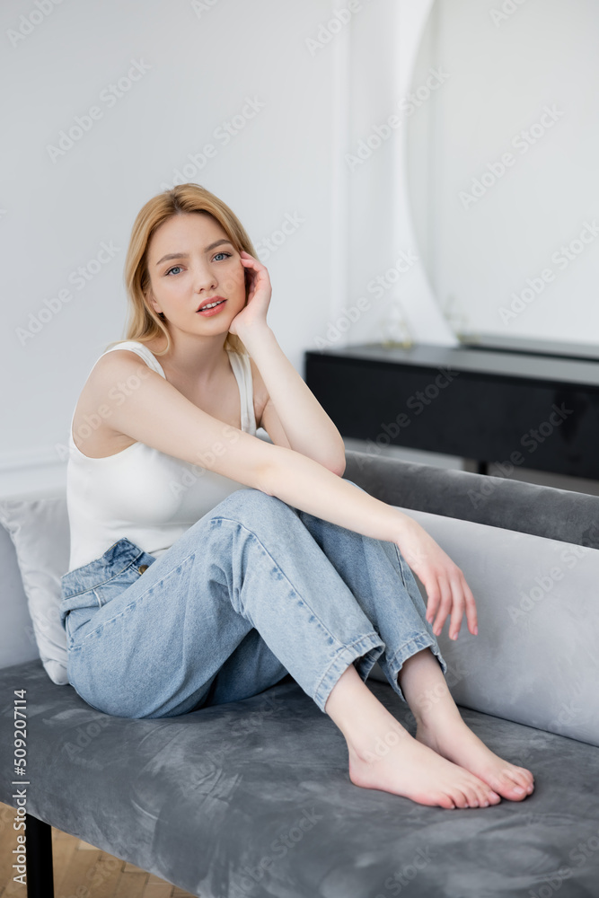Barefoot woman in jeans looking at camera while sitting on couch at home. Barefoot woman in jeans looking at camera while sitting on couch at home.