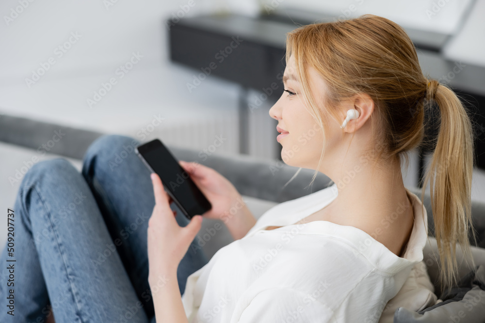 Side view of pretty blonde woman in earphone holding blurred smartphone at home.