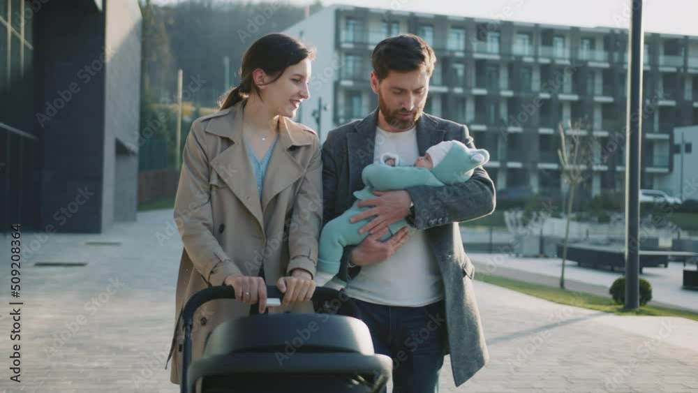 Stylish young family walking with a pram near the lake. Man holding new born baby. Smiling at sunlight. Parenthood. Slow motion Stylish young family walking with a pram near the lake. Man holding new born baby. Smiling at sunlight. Parenthood. Slow motion