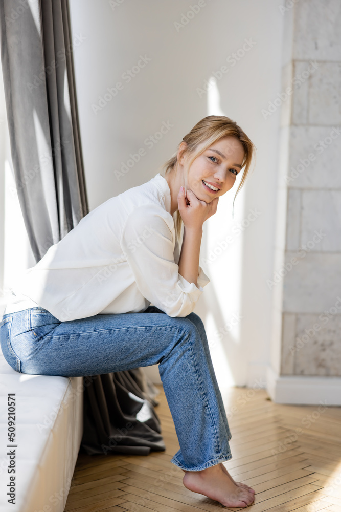 Positive barefoot woman in jeans and blouse looking at camera on windowsill at home. Positive barefoot woman in jeans and blouse looking at camera on windowsill at home.