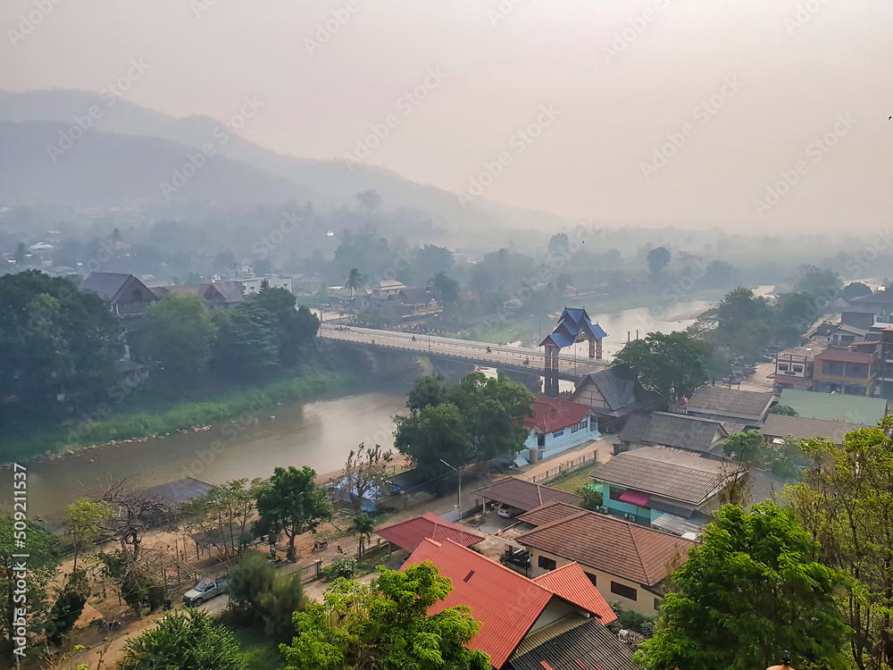 Obraz premium View from the Wat Tha Ton (Thaton) in the north of Chiang Mai province (Thailand) on a misty morning, featuring a bridge with a traditional gate, the river Kok, a village and hills. 