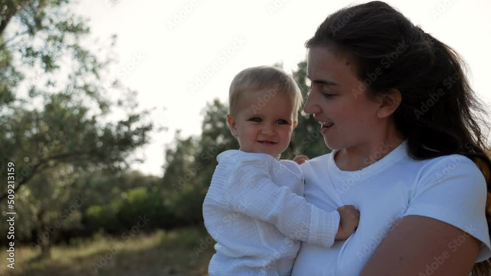 Young mother carries cute son in arms walking in garden with lush trees and greenery. Brunette woman and blond toddler in countryside closeup slow motion Young mother carries cute son in arms walking in garden with lush trees and greenery. Brunette woman and blond toddler in countryside closeup slow motion