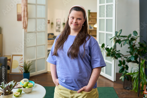 Cheerful girl with Down syndrome wearing blue t-shirt and yellow cotton pants standing in front of camera in spacious living room photo