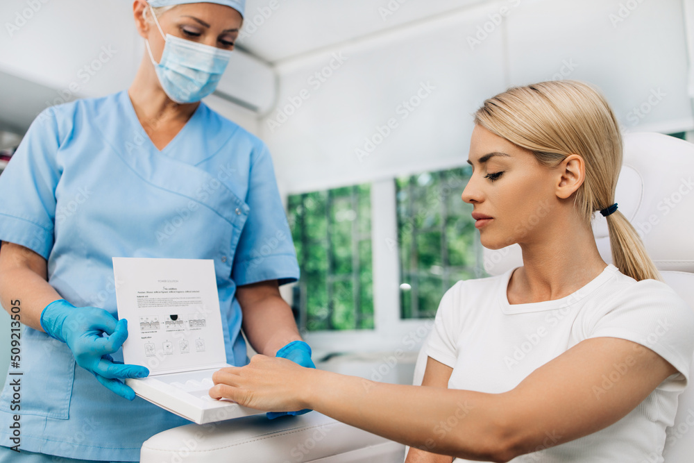Fototapeta premium Beautiful and happy blonde woman at beauty medical clinic. She is sitting and talking with female doctor about face aesthetic treatment.
