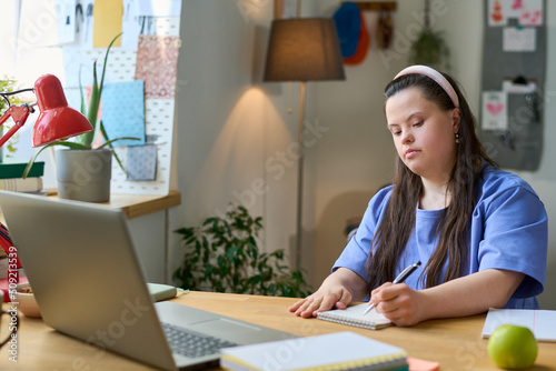 Фотография Diligent girl with Down syndrome making notes in notepad while sitting by desk i