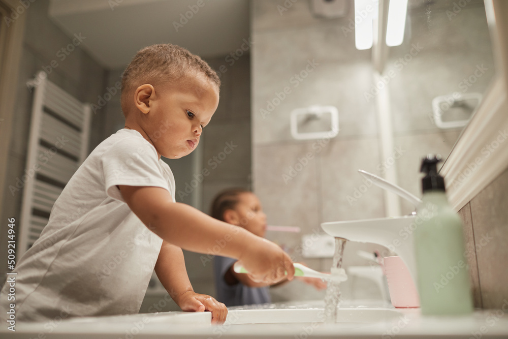 Side view portrait of two African American children in bathroom focus on little toddler boy brushing teeth