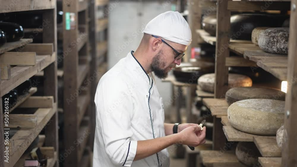 Side view Caucasian cheese maker in eyeglasses smelling tasting delicious aged cheese standing at shelves indoors. Confident concentrated man eating sample from cheese tester in slow motion Side view Caucasian cheese maker in eyeglasses smelling tasting delicious aged cheese standing at shelves indoors. Confident concentrated man eating sample from cheese tester in slow motion