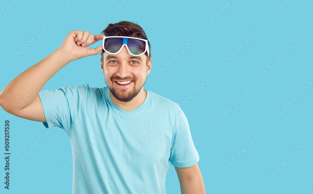 Studio shot of happy handsome man in comfortable summer clothes. Young guy in blue tee shirt standing on solid blue background lifts his sunglasses, looks at camera and smiles. Casual fashion concept Studio shot of happy handsome man in comfortable summer clothes. Young guy in blue tee shirt standing on solid blue background lifts his sunglasses, looks at camera and smiles. Casual fashion concept