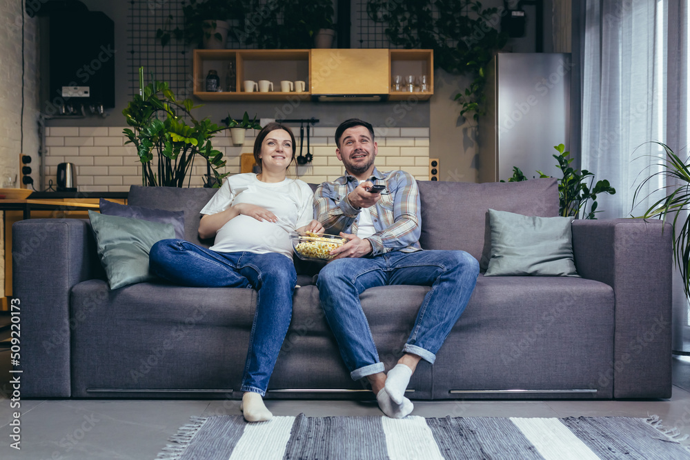 Home cinema. Young family. Pregnant woman and man sitting on sofa at home, watching TV, movie, eating popcorn. Have a good time together Home cinema. Young family. Pregnant woman and man sitting on sofa at home, watching TV, movie, eating popcorn. Have a good time together