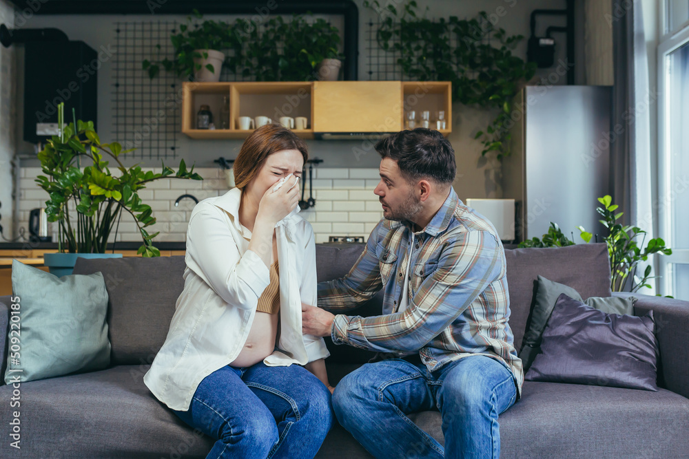 Young pregnant woman crying, wipes tears with a napkin. The man calms her down, supports her. Sitting at home on the couch. Emotional state of a pregnant woman Young pregnant woman crying, wipes tears with a napkin. The man calms her down, supports her. Sitting at home on the couch. Emotional state of a pregnant woman
