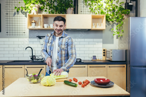 Young handsome man prepares healthy and tasty food in the kitchen at home. Meal and dinner. chops vegetables and fruits. He looks at the camera, smiles