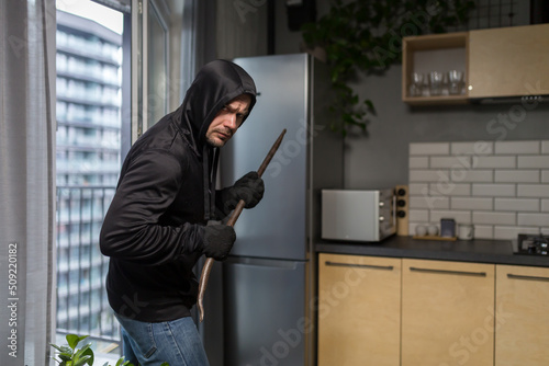 Young male robber in dark clothes carefully and quietly enters through the balcony window into the apartment, house. Holds scrap metal, fittings.