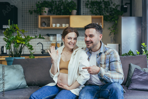 Young family sitting on sofa at home. They talk on a video call. Pregnant woman and man. Woman waving her hand at the camera, smiling. The man points a finger at her belly. Happy