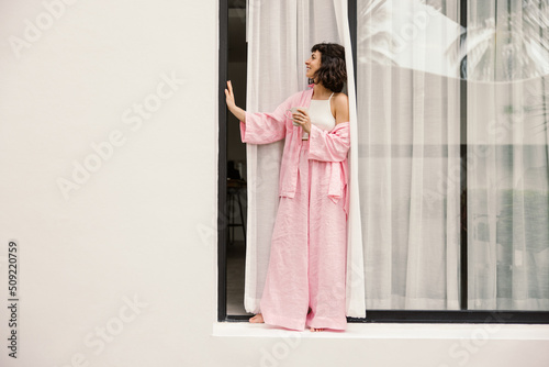 Full-length young caucasian girl stands near open window with cup of strong coffee. Brunette wears white top, pink shirt and pants. Leisure lifestyle and beauty concept.