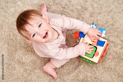 Happy toddler baby boy is playing with a busyboard cube on the nursery floor. Educational toys for children, wooden game panel. A child aged one year photo