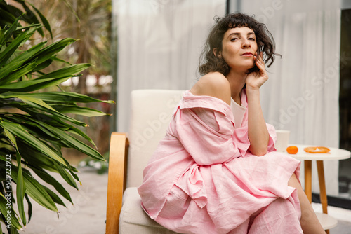 Pretty young caucasian woman looking at camera sitting on armchair on terrace during day. Brunette with short haircut wears pink summer suit. Vacation, happiness and pleasure concept