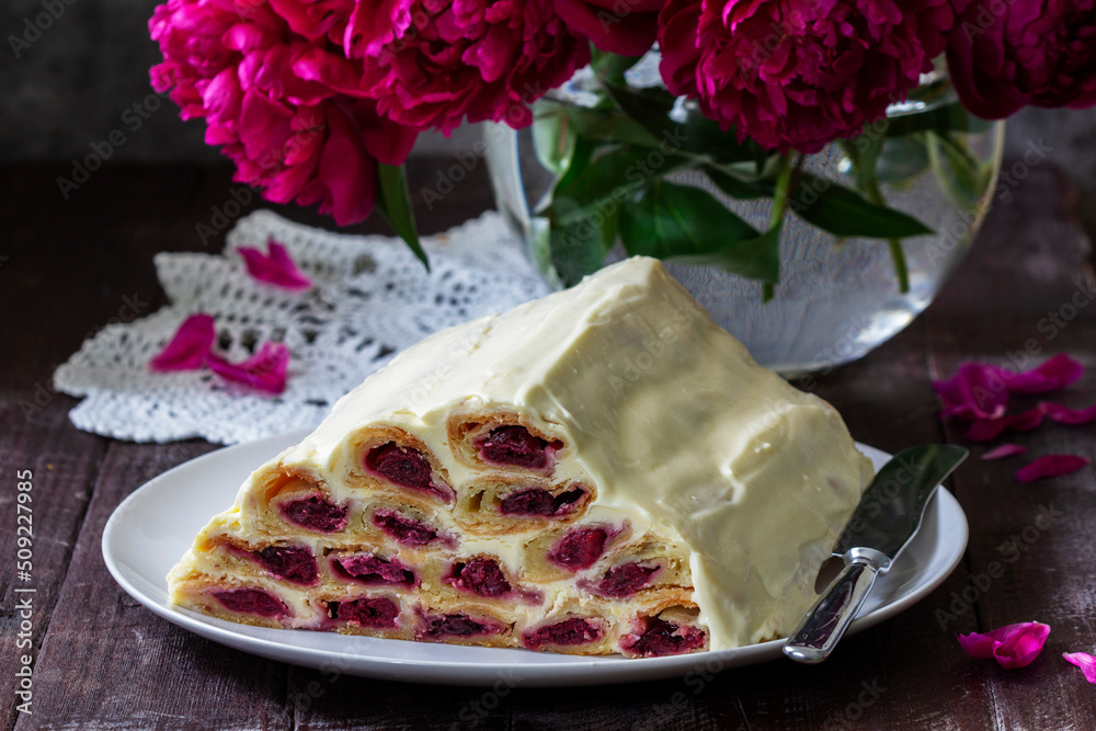 Cake Monastic hut with cherry filling and sour cream, a bouquet of peonies on a wooden table.