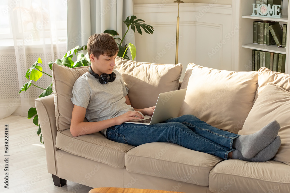 A teenager lying on a beige sofa, in room with plant, wearing black headphones around his neck, looks into a laptop. A teenager lying on a beige sofa, in room with plant, wearing black headphones around his neck, looks into a laptop.