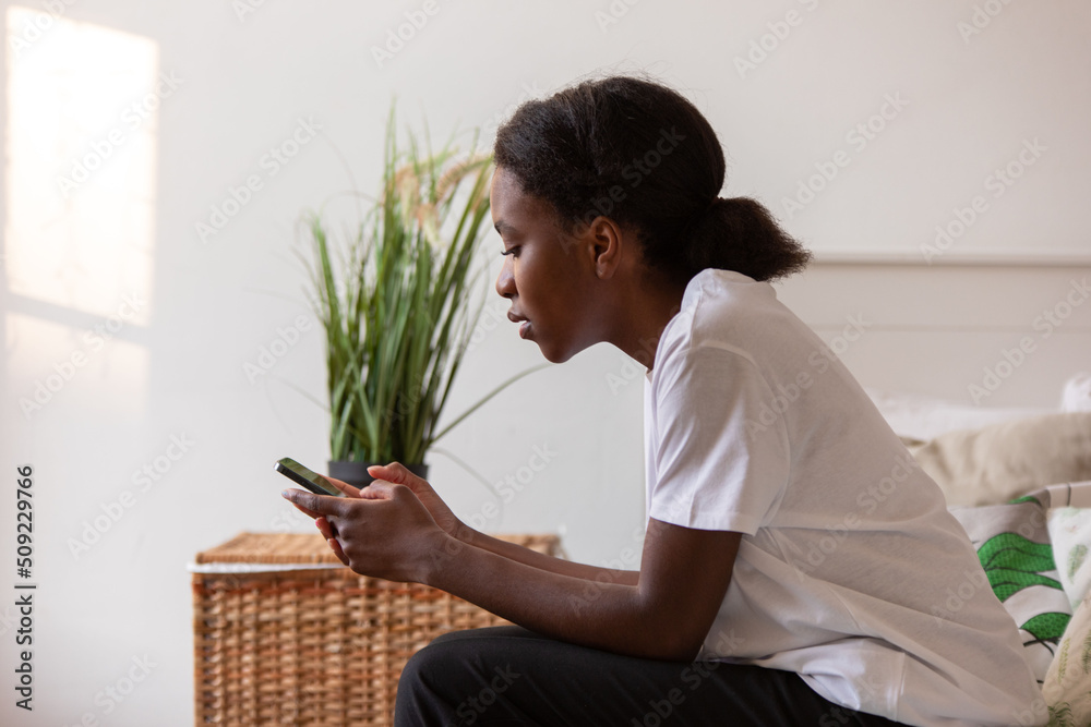Young african american woman sitting on bed using phone.