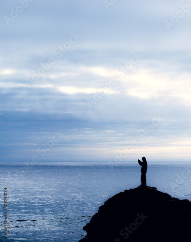 Silueta de hombre con cámara sobre una roca en primer plano con paisaje del mar de playa en día nublado de fondo