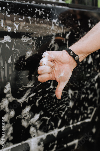 A male professional worker, a washerman, is dissatisfied with his work, showing a thumbs down against the background of a black surface of a car door and white soap suds.