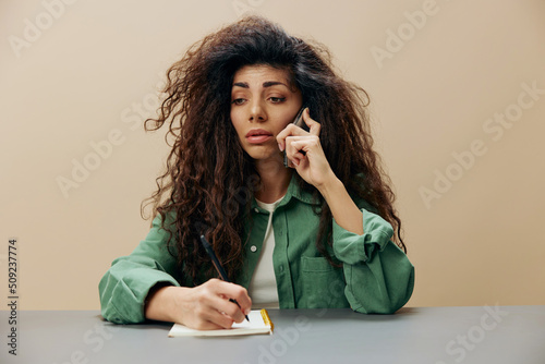 Fotografia Upset unhappy tanned curly Latin lady talking with phone sit at the table write in notebook isolated over pastel beige background look down