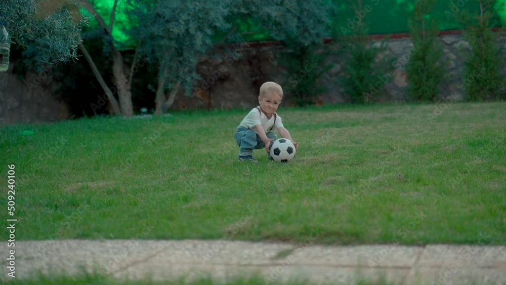 Adorable toddler plays with soccer ball walking on green grass lawn in backyard of country house. Sweet blond boy enjoys sports game at home Adorable toddler plays with soccer ball walking on green grass lawn in backyard of country house. Sweet blond boy enjoys sports game at home