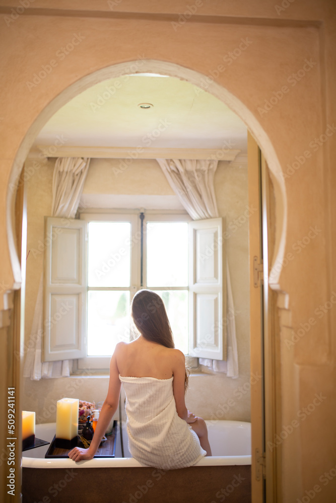 Slim white woman with long hair in the bath room with window. White towel on the body. Morning time. Slim white woman with long hair in the bath room with window. White towel on the body. Morning time.