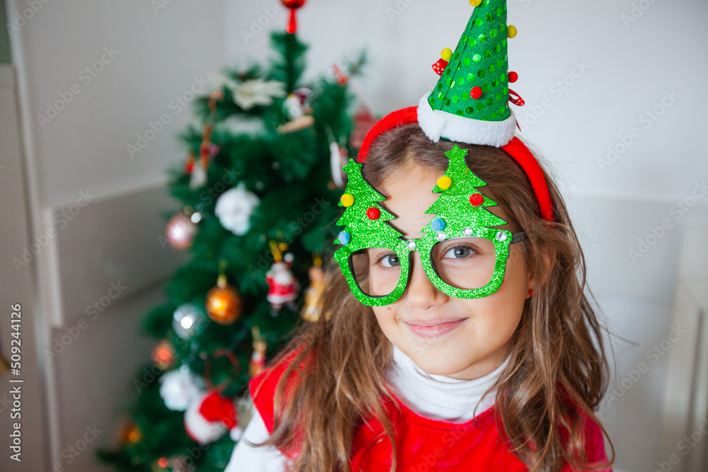 Happy little girl with Santa hat by decorated Christmas tree at home. Child wear fun holidays glasses. Looking at camera. Happy little girl with Santa hat by decorated Christmas tree at home. Child wear fun holidays glasses. Looking at camera.