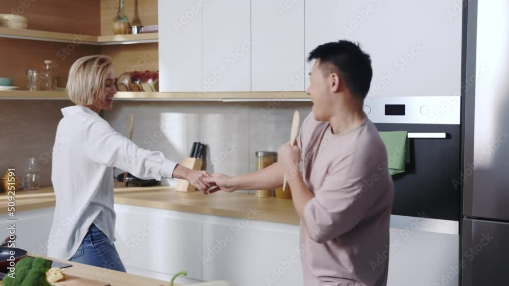Happy multiethnic couple in love dancing and singing in kitchen, using wooden spatulas instead of microphones. Vegetarian. Cheerful beautiful woman and handsome man having fun together at home Happy multiethnic couple in love dancing and singing in kitchen, using wooden spatulas instead of microphones. Vegetarian. Cheerful beautiful woman and handsome man having fun together at home